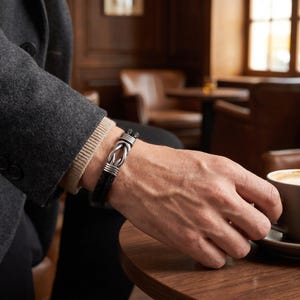 May include: A black leather bracelet with a silver-colored knot design. The bracelet is worn on a person's wrist, with a dark gray jacket and a beige sweater visible in the background. The person is holding a coffee cup.