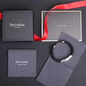 May include: A black leather bracelet with a silver clasp and accents, presented with a gift box, a card that says "THANK YOU", and a red ribbon. The gift box and pouch are branded with the name "Berradas".