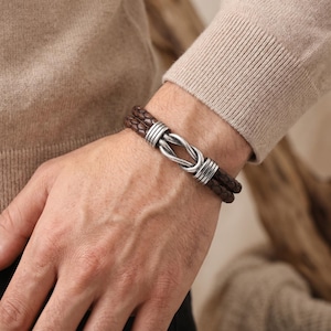 May include: Brown leather bracelet with a braided design and a silver-colored clasp. The bracelet features two strands of braided leather and a decorative clasp. The bracelet is worn on a wrist.
