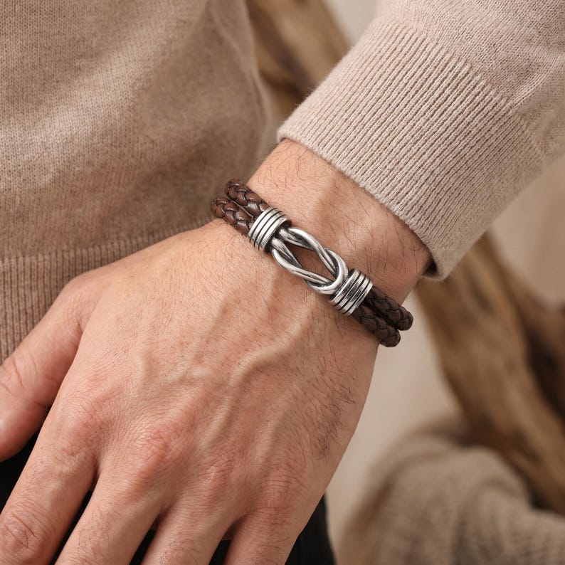 May include: Brown leather bracelet with a braided design and a silver-colored clasp. The bracelet features two strands of braided leather and a decorative clasp. The bracelet is worn on a wrist.