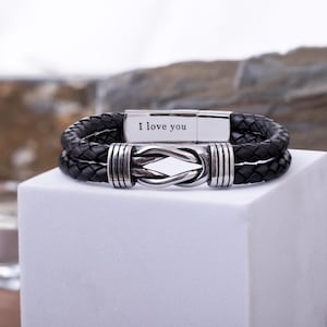 May include: Black leather bracelet with a braided design and a silver clasp. The clasp is engraved with the words "I love you". The bracelet features a knot design in the center.