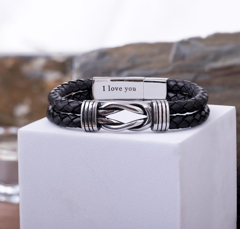 May include: Black leather bracelet with a braided design and a silver clasp. The clasp is engraved with the words "I love you". The bracelet features a knot design in the center.