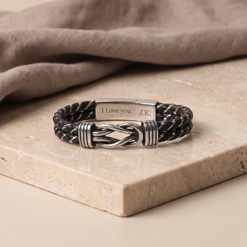 May include: A black and silver braided leather bracelet with a knot design clasp. The bracelet has a magnetic clasp with the engraved text "I Love you J.K." The bracelet is displayed on a light-colored stone surface.