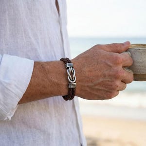 May include: Brown leather bracelet with a silver knot design. The bracelet features braided leather and silver accents. The jewelry is worn on a wrist, with a white shirt in the background.