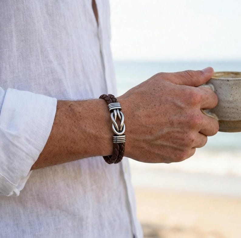 May include: Brown leather bracelet with a silver knot design. The bracelet features braided leather and silver accents. The jewelry is worn on a wrist, with a white shirt in the background.