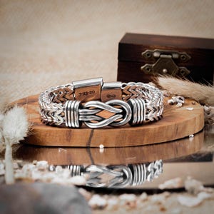 May include: A silver bracelet with a braided chain and a knot-shaped clasp. The clasp has the inscription "7-23-20" and an infinity symbol.