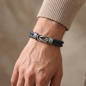 May include: A dark blue leather bracelet with a silver-colored knot design. The bracelet features two braided leather strands and silver accents. The bracelet is worn on a wrist.
