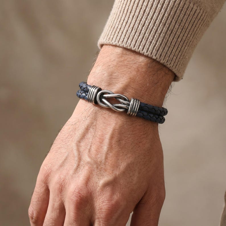 May include: A dark blue leather bracelet with a silver-colored knot design. The bracelet features two braided leather strands and silver accents. The bracelet is worn on a wrist.