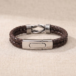May include: A brown leather bracelet with a braided design. The bracelet features a stainless steel clasp and decorative accents. The bracelet is shown on a neutral background.