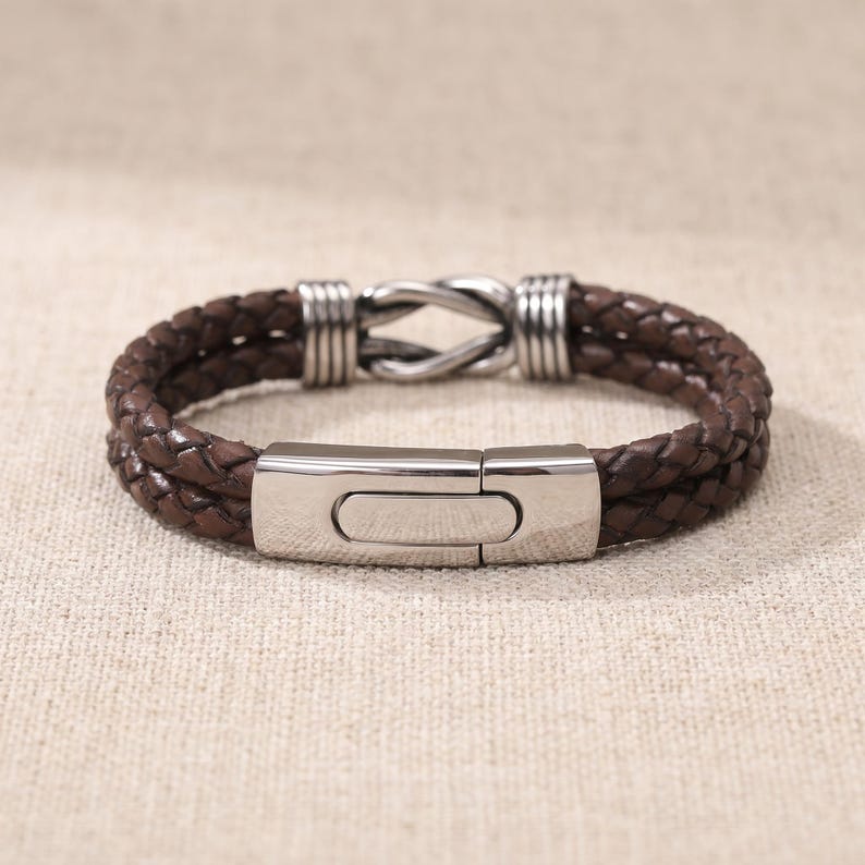 May include: A brown leather bracelet with a braided design. The bracelet features a stainless steel clasp and decorative accents. The bracelet is shown on a neutral background.