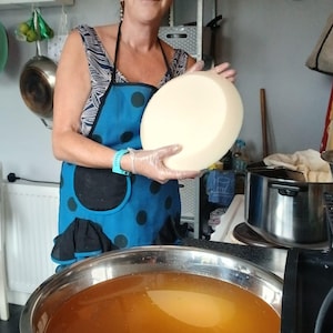 Puede incluir: Una mujer con gafas y un delantal azul sostiene una gran rueda de queso redonda y amarillo pálido. Un gran cuenco de acero inoxidable lleno de líquido ámbar está en primer plano. Ambiente de cocina con ollas y sartenes visibles.