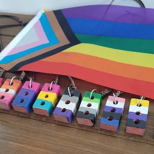 May include: A collection of colorful brick-shaped pendants with various LGBTQ+ pride flag designs. Each pendant is strung on a brown cord. A rainbow flag and a transgender pride flag are also visible.