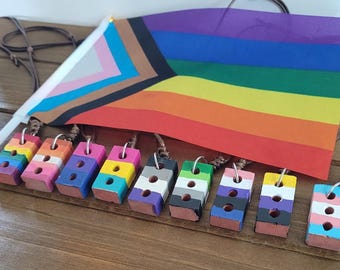 Könnte beinhalten: Eine Sammlung farbenfroher, ziegelsteinförmiger Anhänger mit verschiedenen LGBTQ+-Pride-Flaggen-Designs. Jeder Anhänger ist an einer braunen Kordel befestigt. Eine Regenbogenflagge und eine Transgender-Pride-Flagge sind ebenfalls zu sehen.