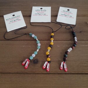 May include: Three beaded necklaces with red ruby slipper charms. Each necklace has a different colored stone: green aventurine, yellow quartz, and bloodstone. The tags for each necklace list the stone type and its properties.
