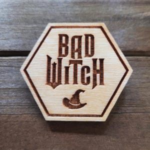 May include: A wooden hexagonal sign with the text "BAD WITCH" in a stylized font. A witch's hat is depicted below the text.