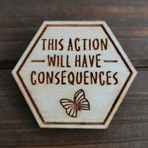 Puede incluir: Placa hexagonal de madera clara con la inscripción grabada "THIS ACTION WILL HAVE CONSEQUENCES" y una ilustración de mariposa. Los detalles grabados son de color marrón oscuro.