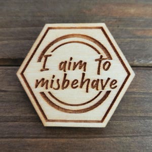 May include: A wooden hexagon-shaped plaque with the phrase "I aim to misbehave" burned into the surface. The text is in a handwritten style, and the plaque has a light wood grain.