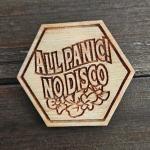 May include: A light-coloured wooden hexagon with the phrase "ALL PANIC! NO DISCO" etched into the surface. Below the text are etched floral designs. The wood has a natural grain and a light brown colour.