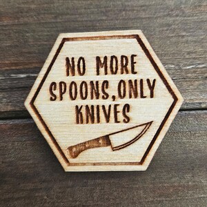 May include: A wooden, hexagonal sign with the phrase "NO MORE SPOONS, ONLY KNIVES" in dark brown lettering. A knife illustration is below the text. The sign is made of light-colored wood with a dark brown border.