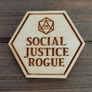 May include: A light-colored wooden hexagon with the words "SOCIAL JUSTICE ROGUE" engraved in a serif font. Above the text is a geometric shape with the number 20 inside. The edges of the hexagon are a darker brown.