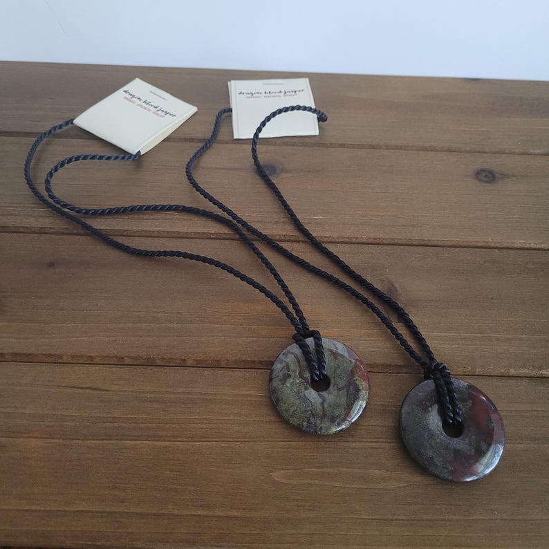 Op de afbeelding: Twee kettingen met ronde hangers van gevlekte groene en rode steen, opgehangen aan zwarte koorden. Elke hanger heeft een centraal gat. Kleine witte labels met tekst zijn aan de koorden bevestigd.
