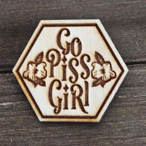 May include: A wooden, hexagonal sign with the phrase "Go Piss Girl" in a decorative font. The sign has floral accents and a light brown color, with darker brown text and details. The sign is likely a decorative item.