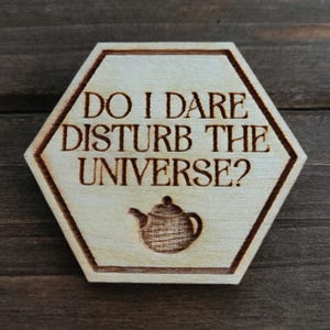May include: A wooden, hexagonal sign featuring the engraved text "DO I DARE DISTURB THE UNIVERSE?" in a serif font, with a teapot illustration beneath. The sign is light brown with a darker brown border.