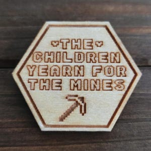 May include: A light-coloured wooden hexagon-shaped item with the text "THE CHILDREN YEARN FOR THE MINES" and a pixelated pickaxe. The text and pickaxe are etched into the wood, creating a contrasting design.