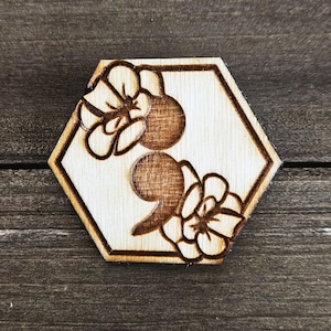 May include: Wooden hexagonal coaster with a brown woodgrain finish. The coaster features a laser-etched design of a semicolon surrounded by two stylised flowers.
