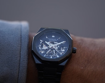 Peut inclure: Une montre squelette noire avec un cadran argenté et un bracelet en métal noir. Le cadran de la montre porte le texte "Thomas & George".