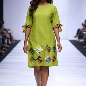 May include: A lime green dress with elbow-length sleeves and a knee-length hemline. The sleeves have patterned cuffs, and the skirt features colourful diamond-shaped appliqués. The model is wearing nude-coloured heels.