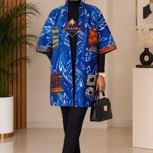 May include: A blue kimono-style jacket with white leaf patterns and contrasting rectangular patches on the sleeves and pockets. The jacket is worn over a black turtleneck and black trousers, paired with white trainers and a black handbag.