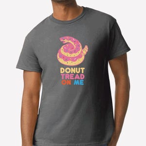 May include: A dark grey t-shirt with a pink donut-shaped rattlesnake with sprinkles. The text "DONUT TREAD ON ME" is printed in rainbow colors below the snake.