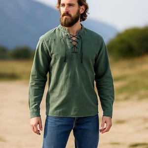 K&ouml;nnte beinhalten: Ein olivgr&uuml;nes Langarmhemd mit Schn&uuml;rkragen und lockerer Passform, kombiniert mit blauen Jeans. Der Hintergrund zeigt eine verschwommene Landschaft mit B&auml;umen und einem Berg.