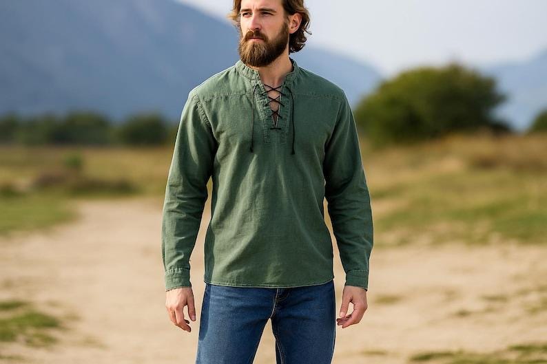 K&ouml;nnte beinhalten: Ein olivgr&uuml;nes Langarmhemd mit Schn&uuml;rkragen und lockerer Passform, kombiniert mit blauen Jeans. Der Hintergrund zeigt eine verschwommene Landschaft mit B&auml;umen und einem Berg.