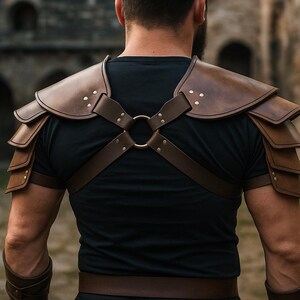 May include: Brown leather shoulder armor with layered plates and decorative rivets. The armor is secured with straps and a central ring, worn over a black shirt. The design suggests a medieval or fantasy theme.