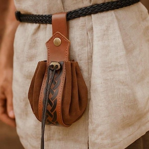 Puede incluir: Una bolsa de cuero y ante marr&oacute;n con un adorno de cuero trenzado, sujeta a una trabilla de cintur&oacute;n de cuero marr&oacute;n. La bolsa se asegura con un cord&oacute;n y un bot&oacute;n de lat&oacute;n. El cintur&oacute;n es negro y trenzado.