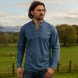 Pode incluir: Uma camisa de ganga de manga comprida com decote com atacadores. A camisa é de cor azul média e tem uma gola clássica. O modelo usa a camisa num campo com um fundo verde.