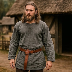 May include: A person wearing a grey tunic and a brown leather belt stands in front of buildings with thatched roofs. The long-sleeved tunic is secured at the waist with a brown belt. The person has long hair and a beard.