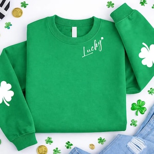 May include: A green sweatshirt with white clover designs on the sleeves and the word "Lucky" in script. The sweatshirt is surrounded by gold coins, shamrocks, and a green hat, all on a white surface. A casual St. Patrick's Day theme.