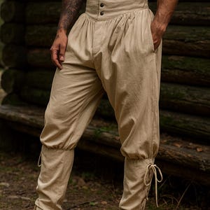 K&ouml;nnte beinhalten: Beige Hose mit hohem Bund, Knopfleiste und gerafften Details. Die Hose hat Taschen und Bindeb&auml;nder an den Kn&ouml;cheln. Der Stoff scheint eine leichte Leinen- oder Baumwollmischung zu sein. Geeignet f&uuml;r historische Nachstellungen oder Kost&uuml;mveranstaltungen.