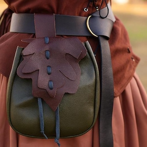 Pode incluir: Uma bolsa de couro artesanal em verde-oliva, com uma aba em forma de folha em castanho escuro. A bolsa está presa a um cinto preto com uma fivela dourada. O design sugere um tema medieval ou de fantasia.