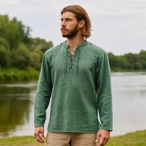 K&ouml;nnte beinhalten: Ein lang&auml;rmeliges, gr&uuml;nes Hemd mit Schn&uuml;rhalsausschnitt. Das Hemd ist aus einem weichen, strukturierten Stoff gefertigt. Das Model steht vor einem Fluss mit B&auml;umen im Hintergrund. Das Hemd ist einfarbig.