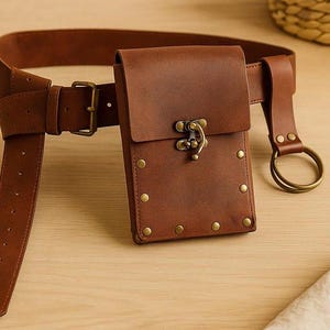 Puede incluir: Un cintur&oacute;n de cuero marr&oacute;n con una funda a juego. El cintur&oacute;n tiene una funda rectangular con cierre de hebilla y remaches de lat&oacute;n. Un anillo de lat&oacute;n est&aacute; unido al cintur&oacute;n. El cintur&oacute;n est&aacute; sobre una superficie de madera.