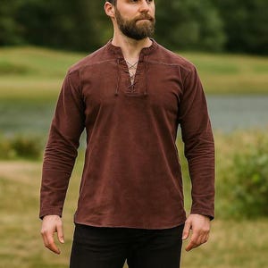 K&ouml;nnte beinhalten: Ein lang&auml;rmeliges, braunes Hemd mit Schn&uuml;rhalsausschnitt. Das Hemd hat ein strukturiertes Aussehen und wird mit einer schwarzen Hose kombiniert. Das Bild wurde im Freien mit einem verschwommenen Hintergrund aus gr&uuml;nem Gras und B&auml;umen aufgenommen.