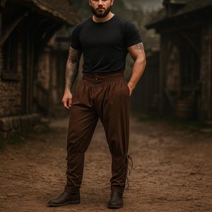 K&ouml;nnte beinhalten: Eine Person tr&auml;gt ein schwarzes T-Shirt und eine braune Hose mit hohem Bund und Knopfverschluss. Die Hose hat eine lockere Passform und ist an den Kn&ouml;cheln gebunden. Die Person tr&auml;gt braune Stiefel. Der Hintergrund ist ein mittelalterliches Dorf.