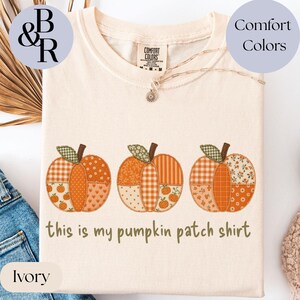 May include: An ivory short-sleeve t-shirt featuring three pumpkins with patchwork patterns in shades of orange, cream, and brown. The text "this is my pumpkin patch shirt" is printed in brown below the pumpkins. The shirt has a Comfort Colors label.