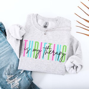 Puede incluir: Una sudadera gris con el texto "Thrifting is my therapy" en rayas verticales de colores. El texto es negro y las rayas son rosa, azul, verde y azul claro.
