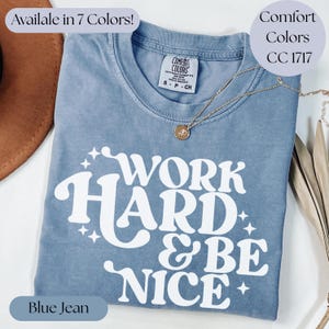 May include: Blue jean colored t-shirt with the words "WORK HARD & BE NICE" in white. The shirt has a crew neck and a small tag on the collar. A gold necklace with a pendant is draped on the shirt. The shirt is available in 7 colors.