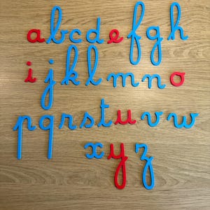 Peut inclure: Un ensemble de lettres de l'alphabet cursives minuscules en bleu et rouge. Les lettres sont disposées sur une surface en bois. Les lettres a, e, i, o, x, y sont rouges. Les lettres restantes sont bleues.